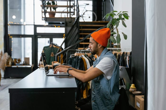 Fashion store employee sitting at counter typing on laptop with clothing racks in background.