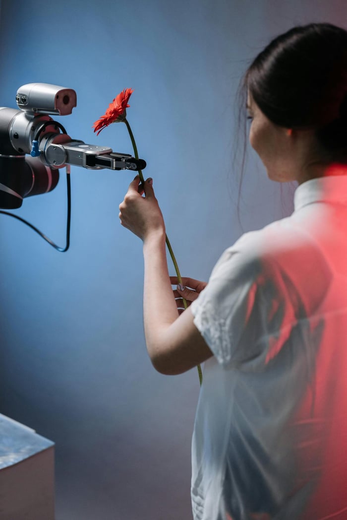 A robotic arm delicately holds a red flower as a woman interacts with it.